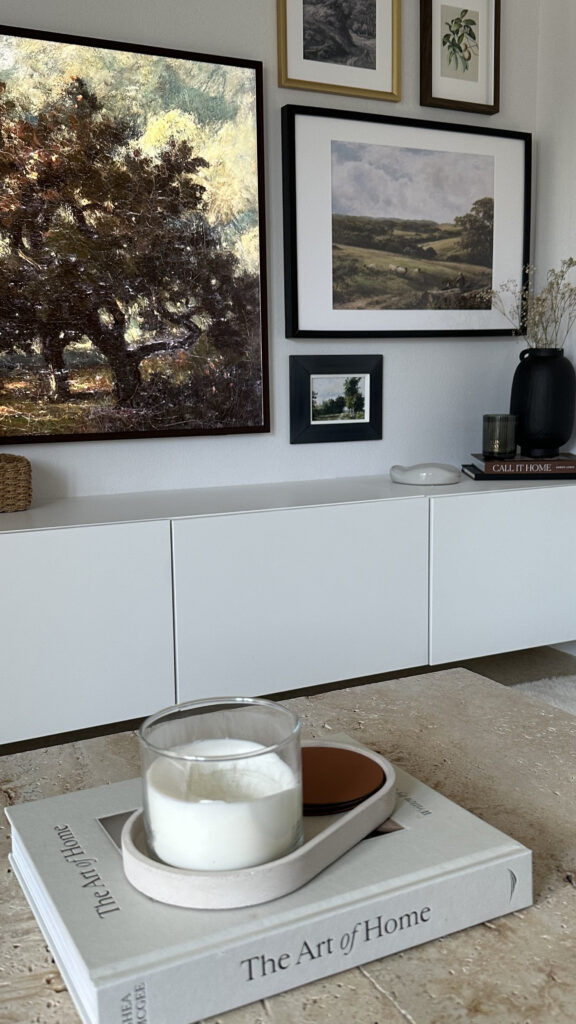 Photo de décoration d'intérieur, en premier plan une table basse en travertin avec un livre de décoration, un plateau effet pierre, une bougie blanche et des dessous de verre. En second plan, un meuble tv blanc de chez Ikea décoré avec des livres, un vase noir garni de fleurs séchées et une bougie. On peut voir sur le mur une tv Samsung The Frame et un mur de cadres