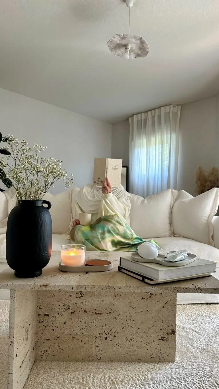 Photo avec en premier plan une table basse en travertin DIY avec des livres, un vase, une bougie et quelques décos. En arrière plan, sur la canapé, une femme qui tient un livre devant son visage